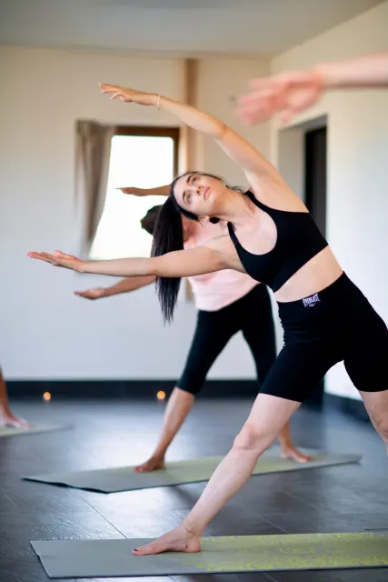 A woman reaches both arms sideways into a standing side stretch while other participants follow on their mats.