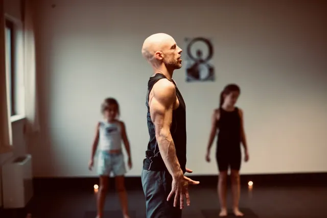 A man stands upright in profile with open hands beside his hips, while two children stand behind him in a studio.
