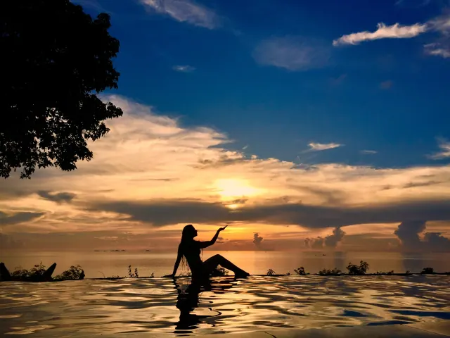 A seated silhouette reaches toward a golden sunset from the edge of an infinity pool, with dark trees framing the sky.