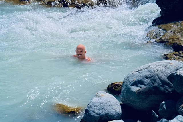 A smiling man swims in pale turquoise river water below foaming rapids, surrounded by dark rocks in an alpine valley.