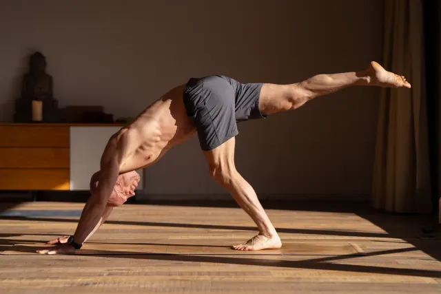 A man holds three-legged downward-facing dog on a mat, lit by low sunlight across the wooden studio floor.