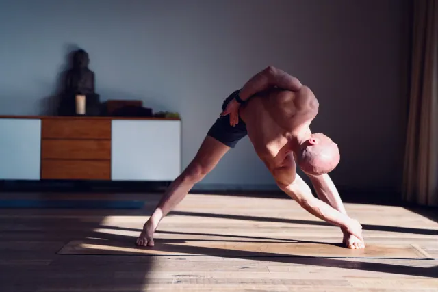 A man twists into a wide-legged standing yoga pose as strong sunlight casts long shadows across the wooden studio floor.