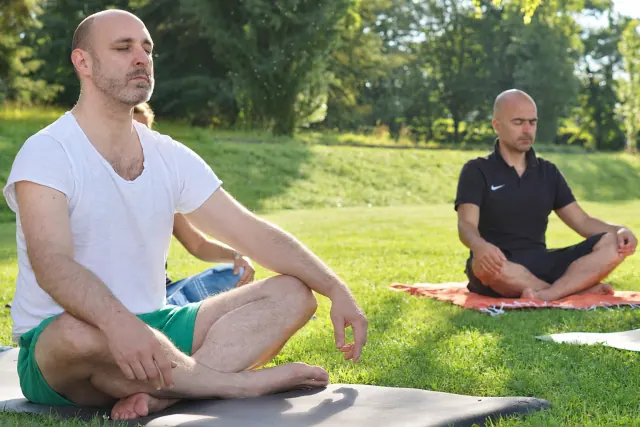 Two men sit cross-legged with their eyes closed on yoga mats in a sunlit park surrounded by green grass and trees.