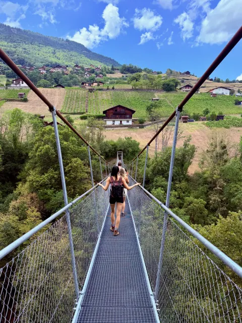 A person walks away across a narrow suspension bridge toward a green hillside dotted with wooden chalets.