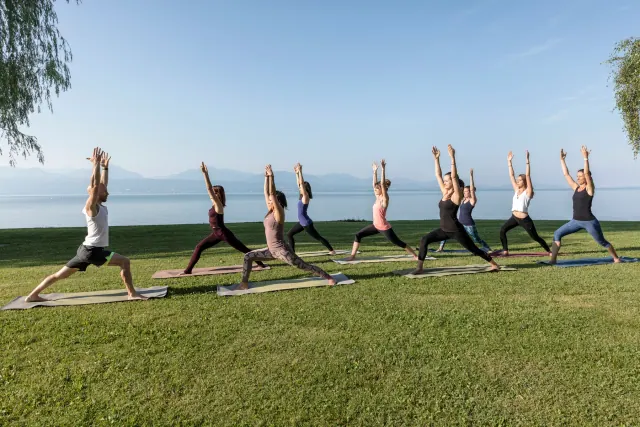 A yoga group holds Warrior I with arms overhead on a sunny lawn beside a lake, with mountains in the distance.