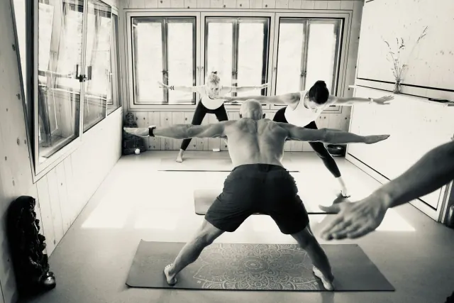 A black-and-white view of a yoga class holding Warrior II with arms extended in a window-lined wooden room.