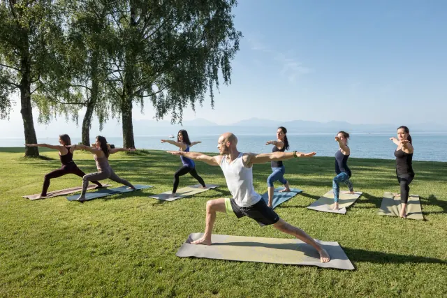 A teacher leads a lakeside yoga group in Warrior II, with arms outstretched beneath trees on a sunny lawn.