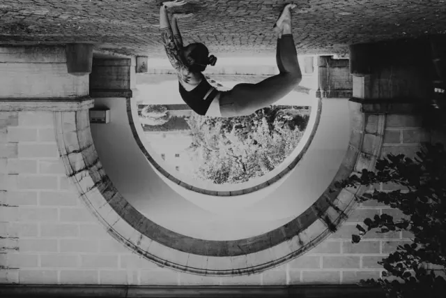 An upside-down black-and-white composition shows a woman holding wheel pose beneath a broad stone archway.