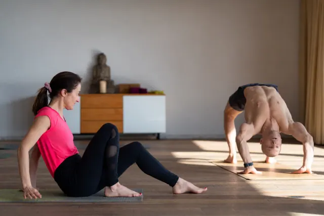 A man holds wheel pose on a mat in a sunlit studio while a seated woman watches from the foreground.