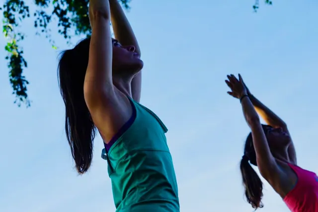 Two women stretch their arms overhead during outdoor yoga, viewed from below against a clear blue sky.