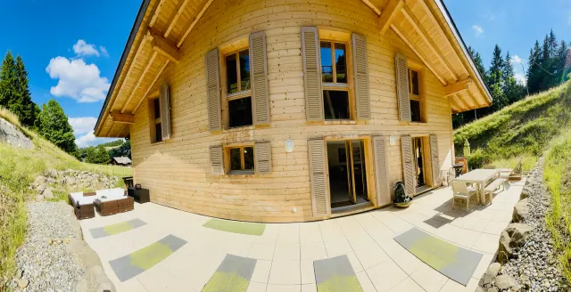 Yoga mats are laid out on a sunny terrace beside a wooden chalet, with grassy slopes and trees around it.