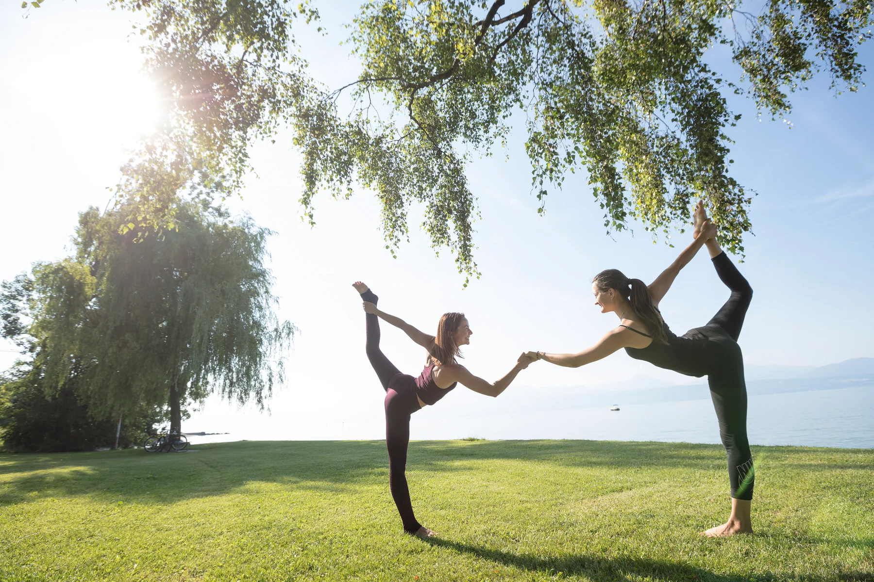 Deux pratiquantes tiennent la posture du danseur au bord du lac, chacune tenant son pied arrière tandis que leurs mains libres se rejoignent pour s’appuyer.