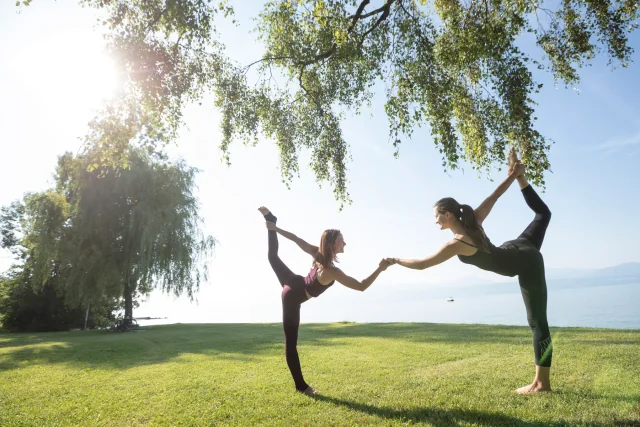 Deux pratiquantes tiennent la posture du danseur au bord du lac, chacune tenant son pied arrière tandis que leurs mains libres se rejoignent pour s’appuyer.