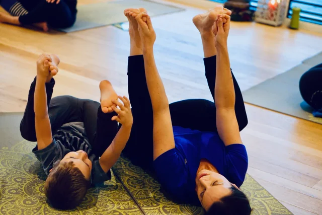 Un adulte et un enfant sont allongés dans la posture du bébé heureux, tenant leurs pieds avec les genoux ouverts de chaque côté du buste.
