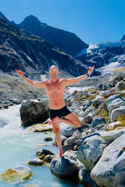 Felix tient la posture de l’arbre parmi des rochers en montagne, un pied contre la jambe d’appui et les bras ouverts.