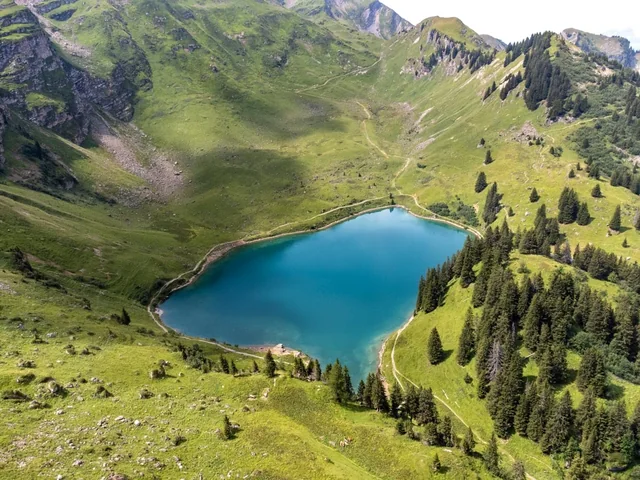 An aerial view shows a deep blue alpine lake nestled in a grassy mountain basin, bordered by rocky slopes and evergreen trees.