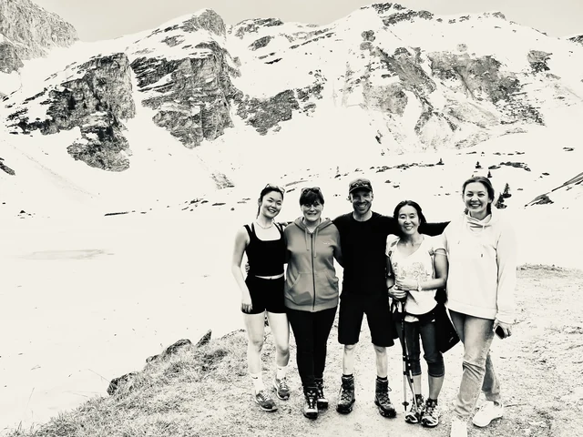 Five people stand together beside an alpine lake and snowy mountains in a black-and-white group portrait.
