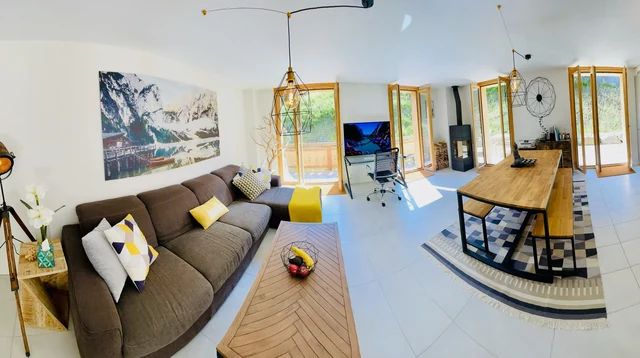 A bright chalet living room has a long dining table, a large sofa and open doors leading toward a sunny terrace.