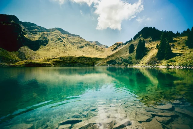 Clear turquoise lake water reveals pale rocks near the shore and reflects green alpine slopes beneath scattered clouds.