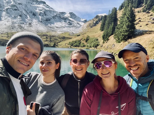 Five smiling hikers pose for a close group selfie beside an alpine lake, with evergreen trees and snowy slopes behind them.