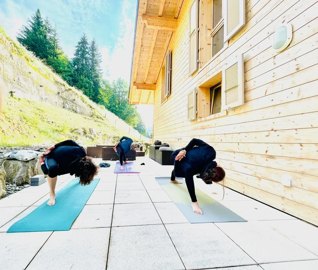 Three women fold forward over bent knees with their hands clasped behind them on a sunny chalet terrace.