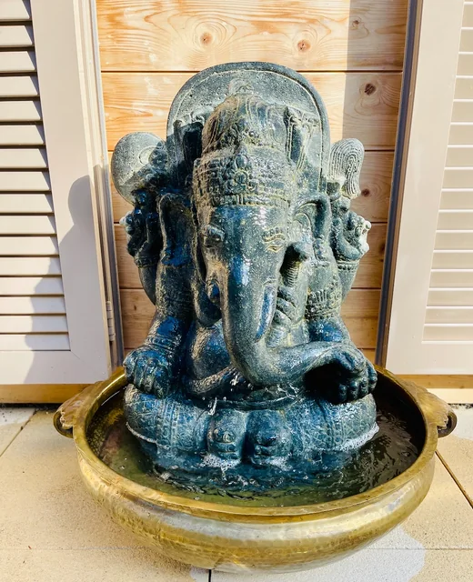 An elephant-headed Ganesha statue sits in a round brass-coloured fountain basin against a wooden wall.