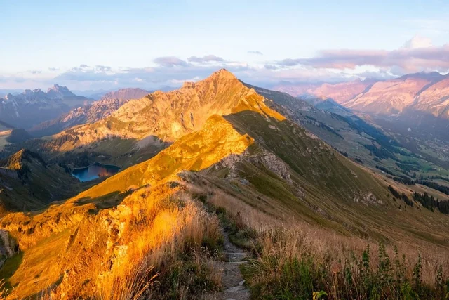 Golden light illuminates a rugged mountain ridge and a narrow hiking trail, with a small lake and layered peaks beyond.