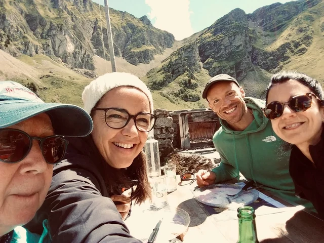 Four smiling hikers gather around an outdoor terrace table for a selfie, with steep rocky mountain walls behind them.