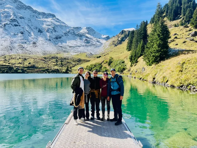 Five hikers stand together on a small jetty over a turquoise alpine lake, framed by snowy peaks and sunlit green slopes.