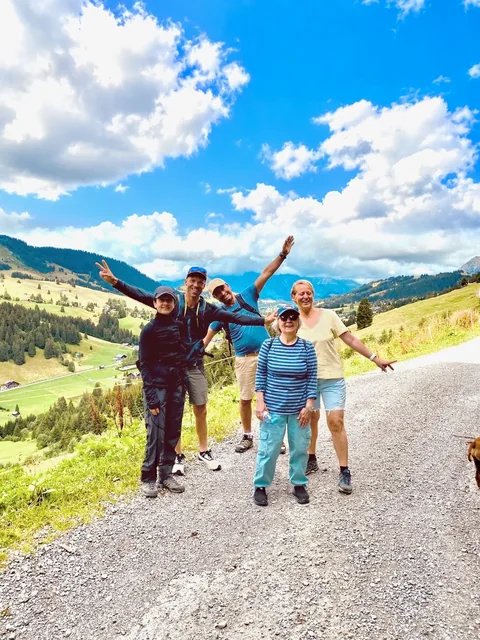 A hiking group smiles on a gravel mountain trail, with raised arms, green alpine slopes and white clouds behind them.