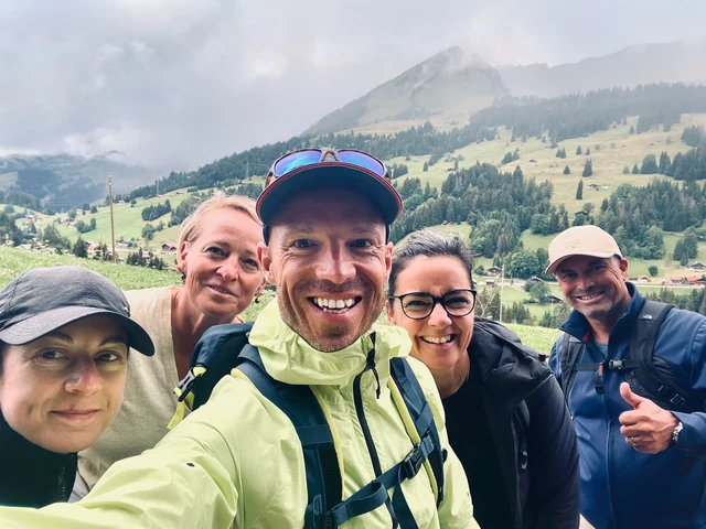 Five smiling hikers pose for a close group selfie, with green alpine meadows and cloud-covered mountains behind them.