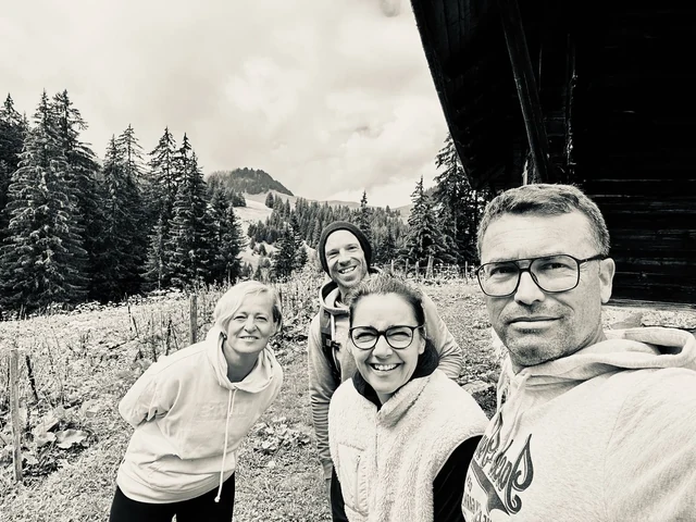 Four hikers smile for a black-and-white selfie beside a wooden chalet, with snowy trees and mountain slopes behind them.