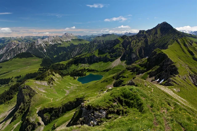 A narrow trail follows a green mountain ridge above a small blue lake, with layered rocky peaks stretching into the distance.