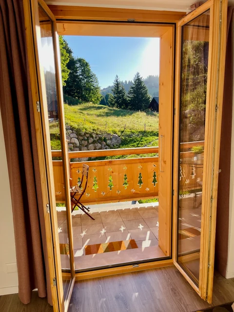 Open wooden balcony doors frame a sunlit alpine meadow and evergreen trees beyond a carved chalet railing.