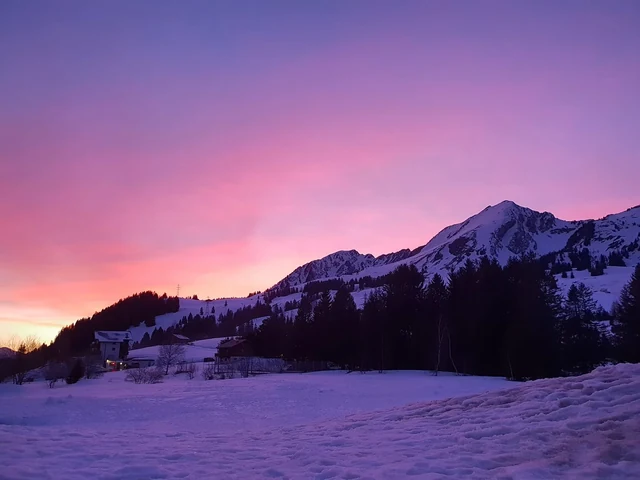 A vivid pink and purple sky hangs above a snowy mountain landscape, with dark evergreen trees and scattered alpine buildings.