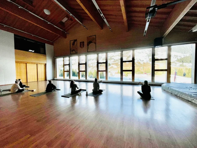 Participants sit quietly on yoga mats in a spacious wooden hall, with green alpine scenery visible through tall windows.