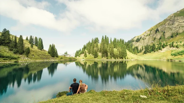 Two people sit together on a grassy lakeshore, looking across calm water that reflects forested slopes and rocky mountains.