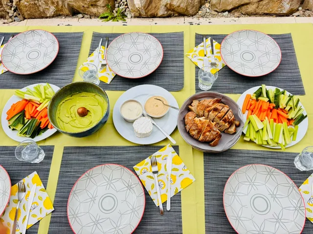 An outdoor dining table is set with patterned plates, raw vegetable sticks, dips and sliced food arranged for a shared meal.