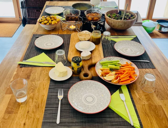 A wooden dining table is set with plates, glasses, bowls of food and colourful sliced vegetables for a shared meal.