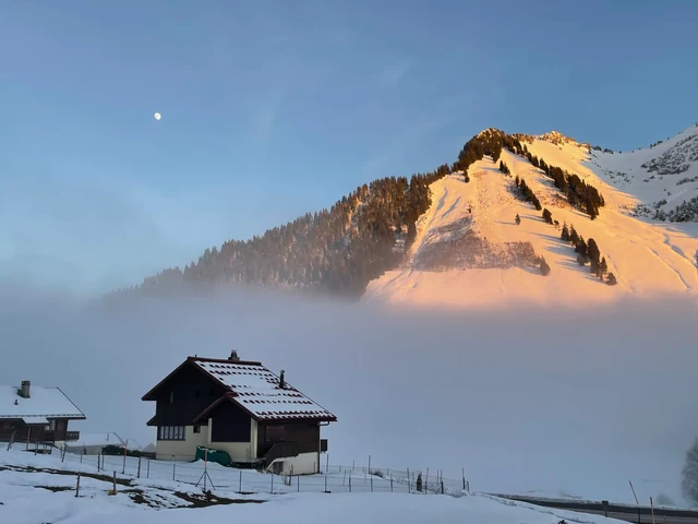 A chalet sits in a snow-covered meadow below a golden mountain peak, with mist filling the valley beneath a pale moon.