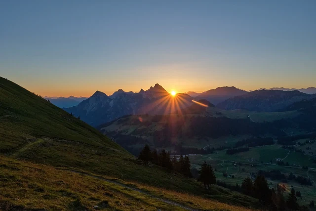 The low sun forms a bright starburst above a mountain ridge, casting golden light across a grassy slope and the valley below.