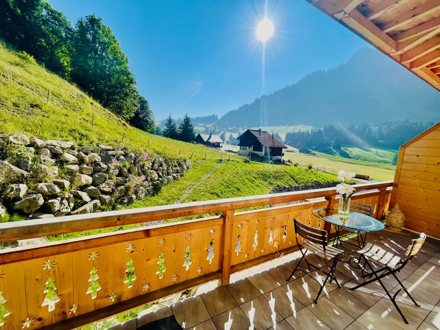 A wooden chalet balcony with a small table and chairs overlooks a sunny green meadow and distant mountains.