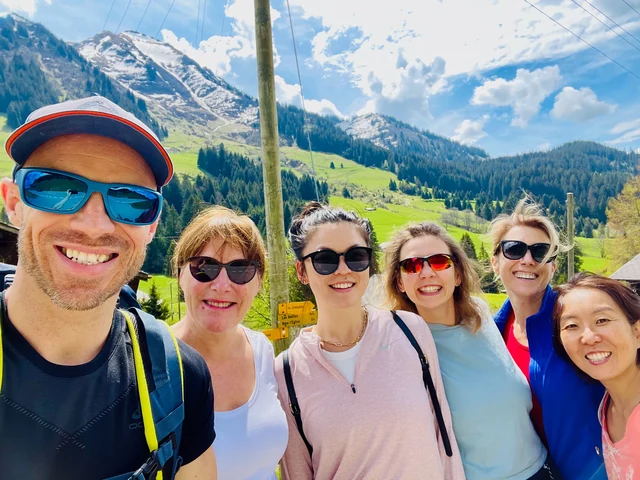 A smiling group poses for a hiking selfie in sunglasses, with green meadows and snowy mountain peaks behind them.