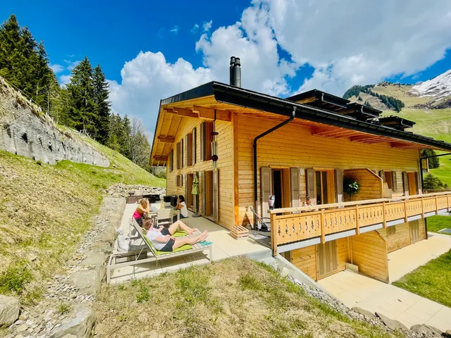 People relax on sun loungers beside a wooden alpine chalet, with a balcony, grassy hillside and blue sky behind them.