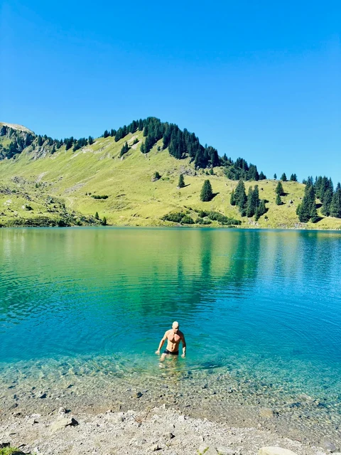 A swimmer stands in clear turquoise water near the shore of an alpine lake, framed by green slopes and a vivid blue sky.