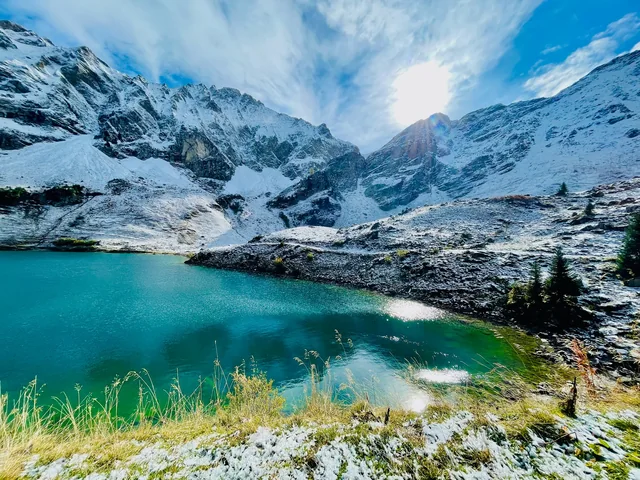 A turquoise alpine lake reflects snowy mountains beneath a bright sky, with frosted grass lining the shore.