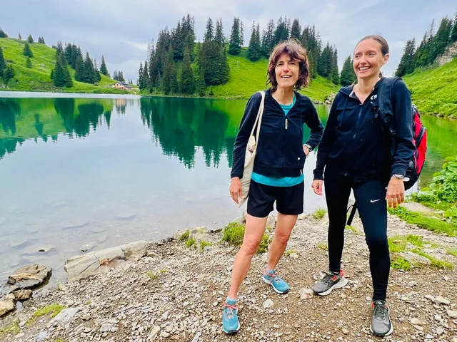 Two smiling hikers stand beside a green alpine lake, with grassy slopes, evergreen trees and their reflections behind them.