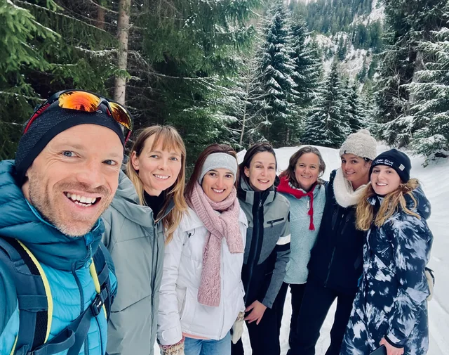A smiling hiking group poses for a selfie in winter clothing, with snow-covered evergreen trees behind them.