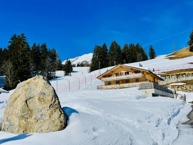 A snowy path passes a large boulder toward a wooden chalet, with evergreen trees and ski slopes beneath a blue sky.