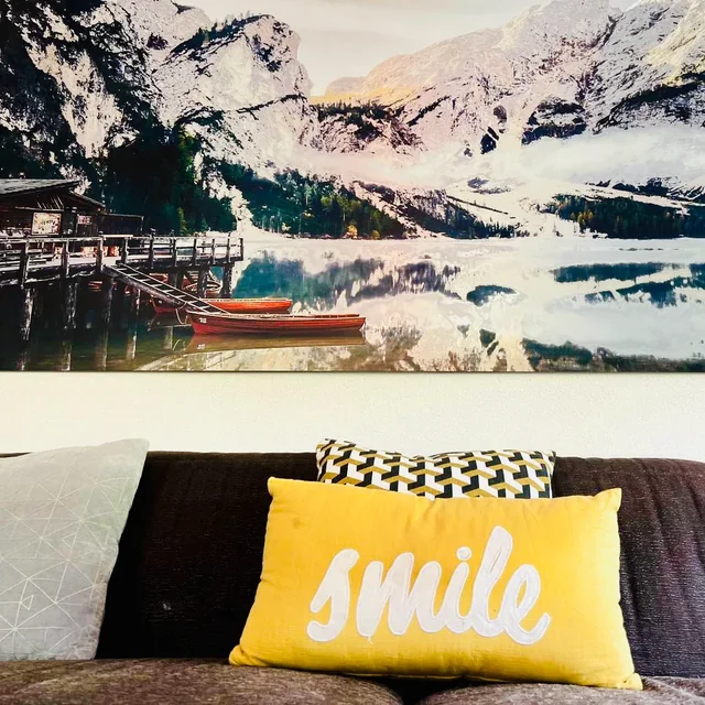 A yellow cushion reading smile rests on a dark sofa beneath a large wall picture of a mountain lake and snow-covered peaks.